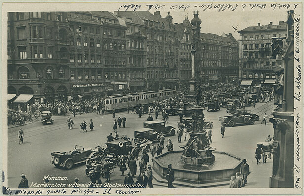 Die Postkarte zeigt den Marienplatz in München während des Glockenspiels. Der Platz ist unübersichtlich: Zu sehen sind parkende wie fahrende Autos, Motorräder, Fahrräder, eine Straßenbahn und vor allem viele Passanten/-innen, die einfach auf der Straße zu laufen scheinen. Straßenschilder, die den Verkehr leiten, oder Linien, die die Straßenführung anzeigen, existieren noch nicht. Bereits in den 1920er-Jahren gab es in Großstädten viele Fahrzeuge, die Motorisierung war in vollem Gang. Angesichts der Unübersichtlichkeit ist eine hohe Unfallrate nicht verwunderlich.